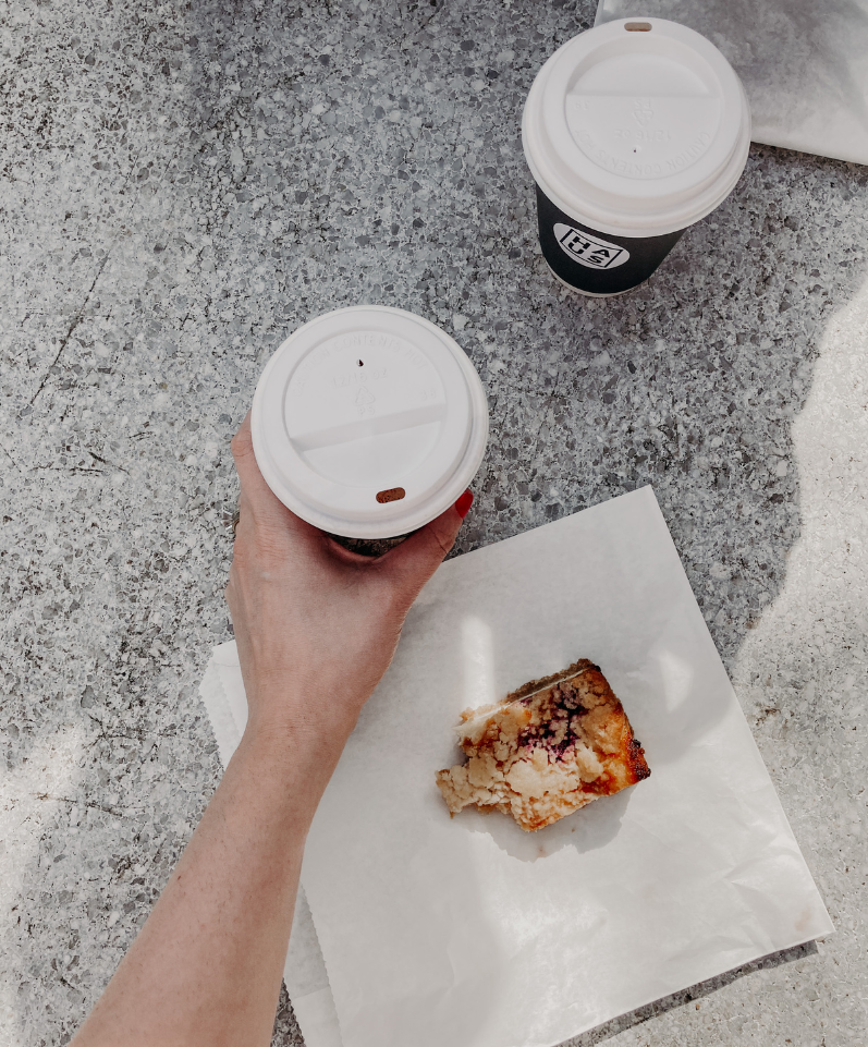 Deux cafés à emporter posés sur une table en pierre, l'un tenu par une main, accompagnés d'une part de gâteau aux fruits sur un papier blanc. Cette scène minimaliste capture l'essence des pauses gourmandes et met en avant l'atmosphère conviviale des meilleurs coffee shops dans le 3ème, des adresses incontournables pour les amateurs de café à Paris.